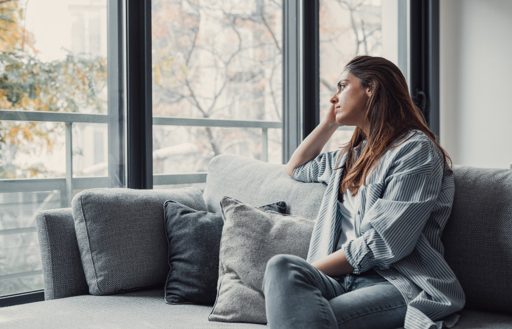 Woman sitting on a couch, looking pensively out a large window. She rests her chin on her hand, with trees visible outside. Martinez, GA