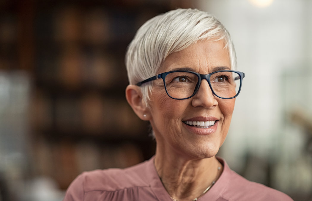 A smiling woman with short white hair and glasses is wearing a pink blouse. She is sitting in a softly lit room with blurred shelves in the background.