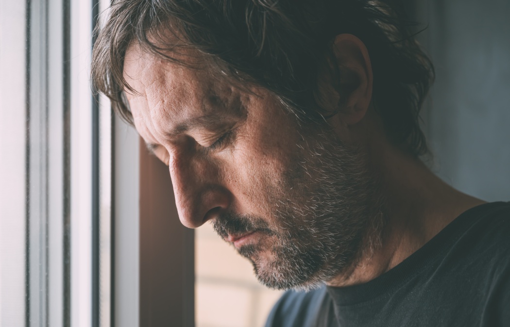 Downhearted man with eyes closed leaning his head close to the apartment window. Depressed unhappy male sad and alone at home. Selective focus. Martinez, GA