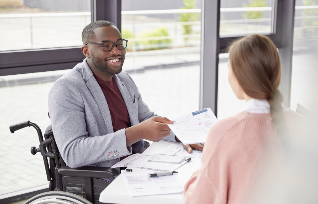 African American male sits at workplace in wheelchair, shares creative ideas and opinions with female worker, demonstrate new business project have informal meeting. Physically handicapped man Martinez, GA
